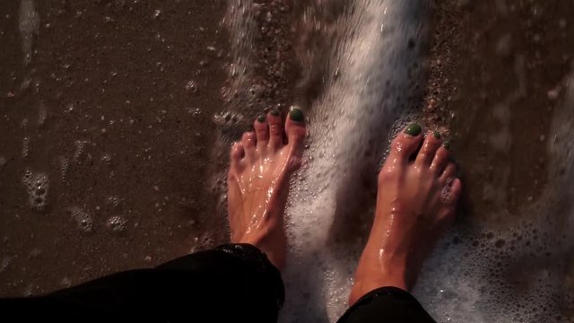 Girl's legs in black jeans on wet sand with shells washes surf, wave with foam