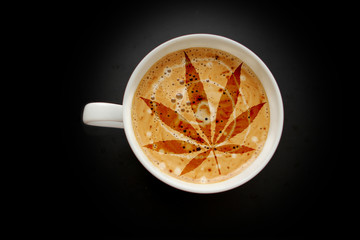 cannabis leaf, marijuana in a white wide cup of coffee with heart crema, cappuccino on a black background with vignette, top view, save space, close-up