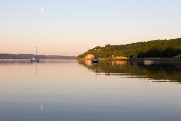 Small anchored sailboats and speedboat in the St. Lawrence River during an beautiful early summer morning with full moon still visible, Cap-Rouge area, Quebec City, Quebec, Canada