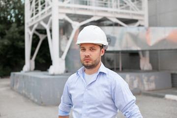 Young handsome male engineer in hardhat standing on the background of water or gas pipes. Concept of boiler station. Pipeline accessories closeup. Heating station manager doing his job outdoor. .