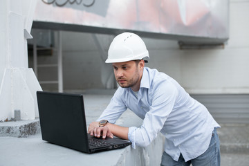 Young engineer in hardhat standing on the background of water or gas pipes station and using laptop. Heating station manager doing his job outdoor.