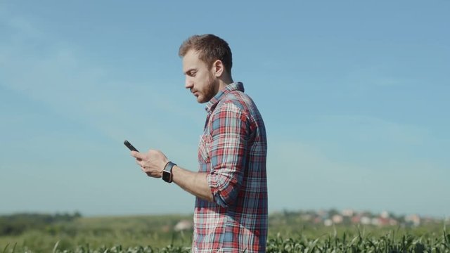 Handsome male traveler walking in the green field in sunlight taking photographs using a smartphone at a farm on summer day.