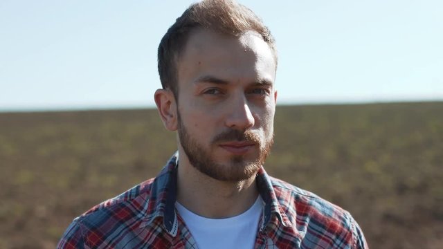 Portrait of handsome happy farmer in shirt enjoying nature in the large rural field. Close-up of smiling young man staying at a farm in sunny weather.