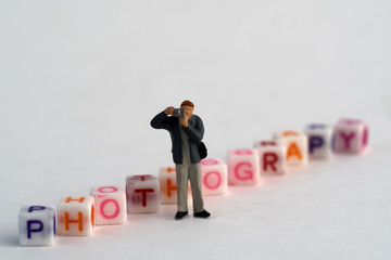Miniature Photographers taking pictures behind a Group Of Letters forming Word Spelling "Photography" on a white surface