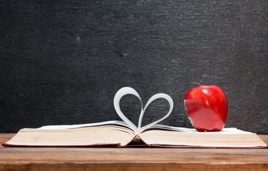 Red apple and book and pages forming heart shape on wooden table