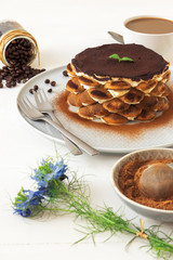 Round tiramisu, homemade no-bake traditional Italian sweet dessert. Table with cake, bowl with cocoa powder, round sponge biscuits, coffee beans and coffee with milk. White background with flowers