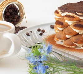 Round tiramisu, homemade no-bake traditional Italian sweet dessert. Blue table with cake, bowl with cocoa powder, round sponge biscuits, coffee beans and coffee with milk. Blue background with flowers