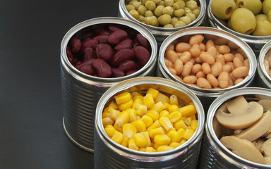 Canned green peas, beans, corn, olives and mushrooms in tin cans. Preserved food on black table.