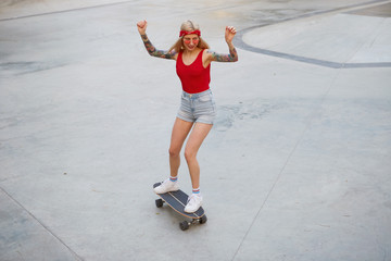 Young cute blonde with tattooed arms in a red T-shirt and denim shorts with a knitted bandana on her head, in red glasses, with hands up, enjoying the day and longboarding in skate park.