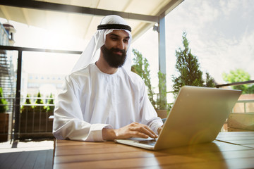 Arab saudi businessman working online with a laptop and tablet in a coffee shop or a cafe with an outdoor terrace in the background. Concept of business, finance, modern technologies, start up.