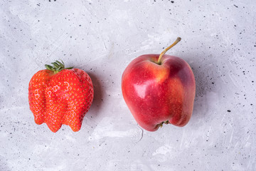 Two ugly fruits: strawberry and apple on grey concrete background. 