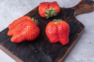 Close-up ugly non-standart ripe red strawberries on burned kitchen board on concrete background. 