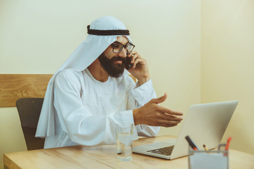 Arab saudi businessman working online with a laptop and tablet in his comfortable cabinet or office. Male model as an enterpreneur. Concept of business, finance, modern technologies, start up, economy