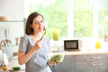 Beautiful young woman eating fresh salad in kitchen