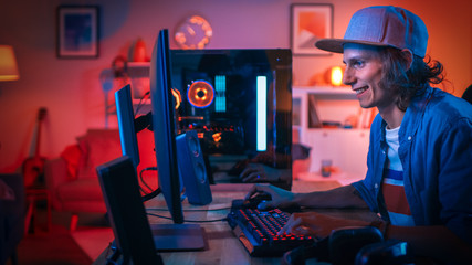 Gamer Playing First-Person Shooter Online Video Game on His Powerful Personal Computer with Colorful Neon Led Lights. Young Man is Wearing a Cap. Living Room Lit with Warm Orange Lamps. Evening. © Gorodenkoff
