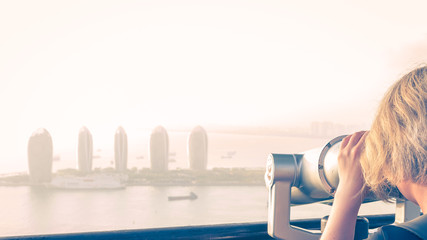 Sanya, Hainan island, China. A girl is watching through binoculars a panorama of Sanya City from a height Luhuitou Park. Sanya Phoenix Island. Sunset, blur, haze.