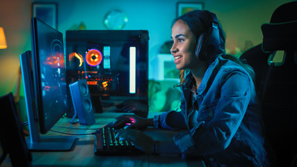 Pretty and Excited Black Gamer Girl in Headphones is Playing First-Person Shooter Online Video Game on Her Computer. Room and PC have Green Neon Led Lights. Cozy Evening at Home.