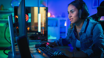 Pretty and Excited Black Blogger Girl in Headphones is Playing First-Person Shooter Online Video Game on Her Computer. Room and PC have Colorful Neon Led Lights. Cozy Evening at Home. © Gorodenkoff