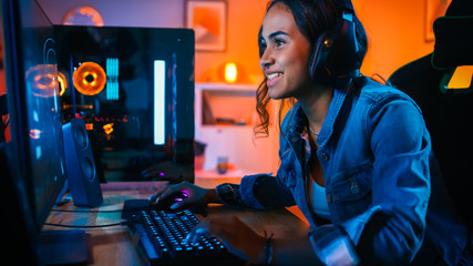 Pretty and Excited Black Gamer Girl in Headphones is Playing First-Person Shooter Online Video Game on Her Computer. Room and PC have Colorful Neon Led Lights. Cozy Evening at Home. - obrazy, fototapety, plakaty