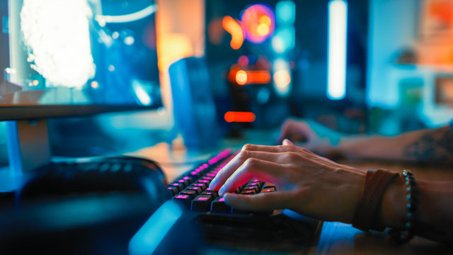 Close-Up Hands Shot Showing a Gamer Using the Keyboard while Playing an Online Shooter Video Game. Keyboard has Pink Neon Lights in Buttons. Gamer is Wearing a Bracelet. Room is Dark.