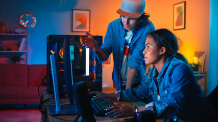 Two Friends or a Couple Discuss Future Strategies on a Computer Game. Pretty Black Girl Shows Her Work to a Young Man and He Shares Creative Ideas. Cozy Room is Lit with Warm and Neon Light.