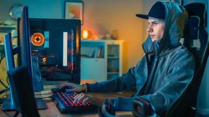 Professional Gamer or Streamer Playing First-Person Shooter Online Video Game on His Cool Personal Computer. Young Man is Wearing a Cap and Hood. Room and PC have Colorful Neon Led Lights.