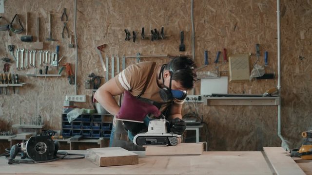 close up of a worker grinds the wood, craftman works in modern furniture factory grinding down sanding wooden part, a lot of tools and equipment on background