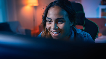 Beautiful Surprised and Excited Young Black Girl Blogger Watching Action Video on a Computer. She Has Dark Hair and Brown Eyes. Screen Adds Reflections to Her Face. Cozy Room is Lit with Warm Light.