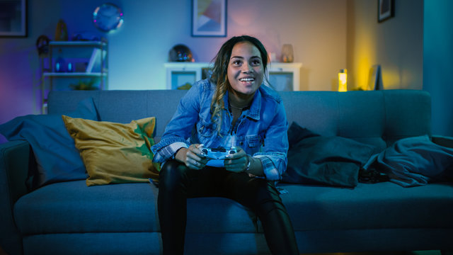 Beautiful Excited Young Black Gamer Girl Sitting On A Couch And Playing Video Games On A Console. She Plays With A Wireless Controller. Cozy Room Is Lit With Warm And Neon Light.