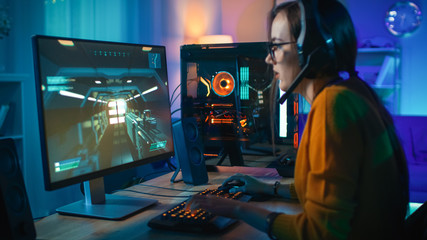 Excited Gamer Girl in Headset with a Mic Playing Online Video Game. Focus is on Her Personal Computer. Room and PC have Colorful Warm Neon Led Lights. Cozy Evening at Home. © Gorodenkoff