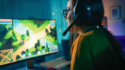 Excited and Concentrated Gamer Girl in Glasses and Headset with a Mic Playing Online Strategy Video Game on Her Personal Computer. Room and PC have Colorful Warm Neon Led Lights. Cozy Evening at Home.