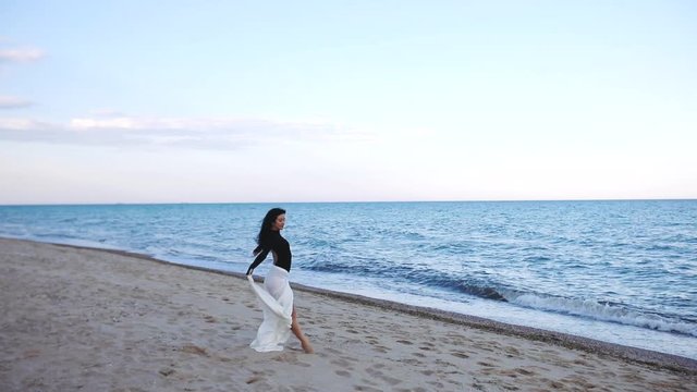 elegant young lady with long white fabric on hips poses on empty sea beach under clear blue sky slow motion