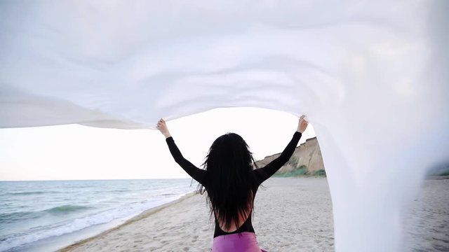long haired woman in pink skirt walks holding sheet of white cloth waved by strong wind over head backside slow motion