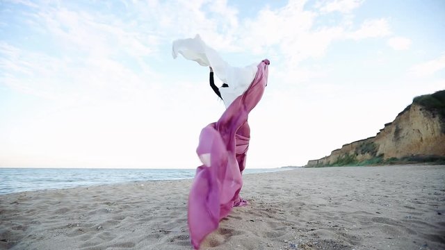 slim long haired girl stands with large chiffon cloth pieces waving in strong summer wind on beach backside slow motion