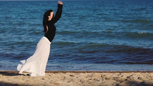 beautiful smiling girl in loose skirt raises leg hands up showing ballet poses on empty beach at azure sea slow motion