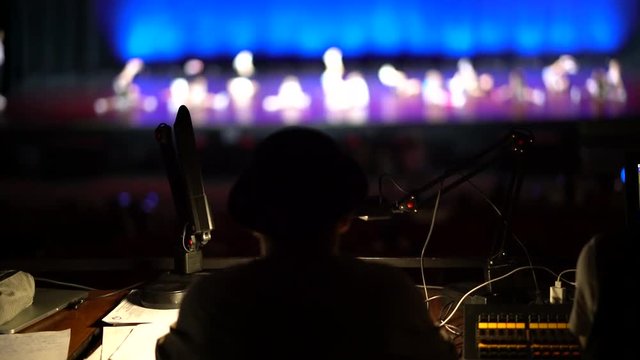 dark silhouette of a man sitting at the operator console in the concert hall on the background of a blurred image of a brightly lit scene with silhouettes of moving people