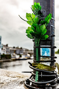 Religious prayer card tied to a metal pole with branches with green plastic leaves on a bridge, a memorial place, rainy day in Ireland