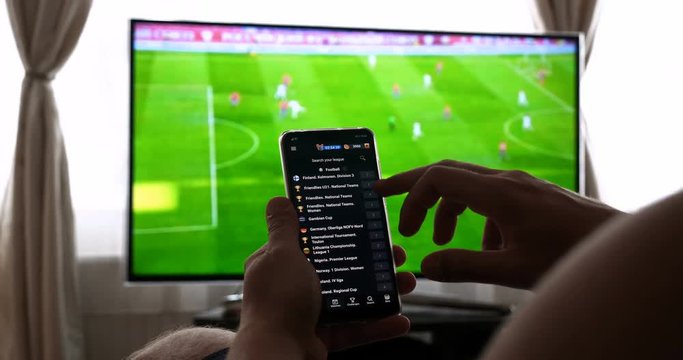Against the background of the TV at home (at the bar), the guy (woman) orders different snacks, pizza and sweets scrolling through internet stores using mobile phones. Concept: Football, Sport,Order.
