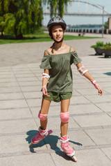 Teenage girl in a helmet learns to ride on roller skates holding a balance or rollerblading and spin at the city's street in sunny summer day. Healthy lifestyle, childhood, hobby, leisure activity.