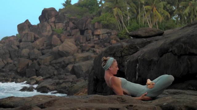 yoga teacher in tracksuit gets into padma matsyasana on large flat rock on sea shore in morning extreme slow motion. Concept yoga meditation and spiritual practices