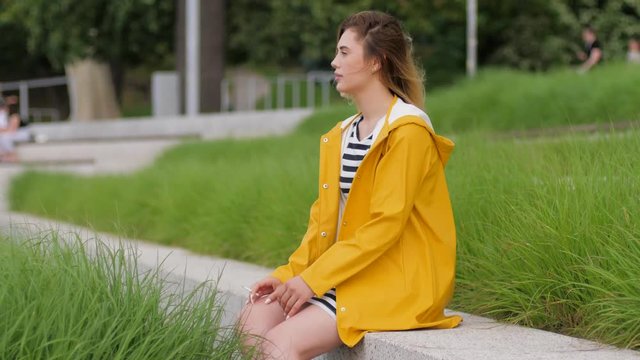 Pretty young girl in yellow rain coat smoking cigarette sitting alone outdoors