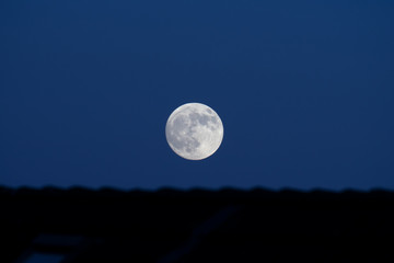 Fototapeta na wymiar Grey full moon in darkening sky above black house roofs