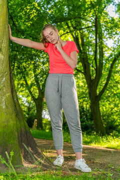 A young jogger leans against a tree and measures her pulse with her hand