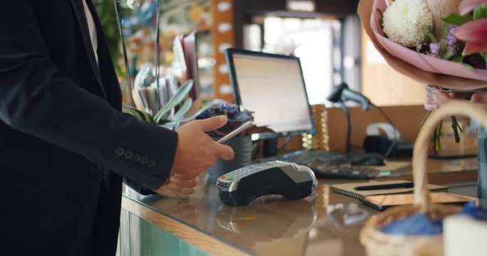 Businessman in suit is buying bouquet of flowers paying online with smartphone holding device making payment in florist's shop. People and gadgets concept.