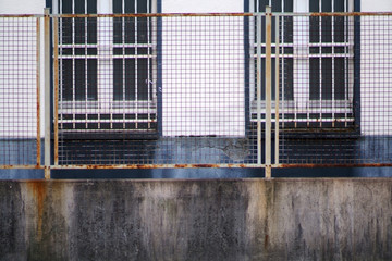 Das Detail eines korrodierenden und rostenden Gitterzaunes vor einer verschlissenen Fassade..