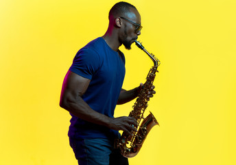 Fototapeta na wymiar Young african-american jazz musician playing the saxophone on yellow studio background in trendy neon light. Concept of music, hobby. Joyful attractive guy improvising. Colorful portrait of artist.