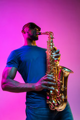 Fototapeta na wymiar Young african-american jazz musician playing the saxophone on pink studio background in trendy neon light. Concept of music, hobby. Joyful attractive guy improvising. Colorful portrait of artist.