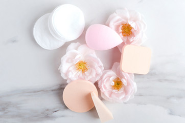 Various types of cosmetic sponges, cotton pads and flowers on the white marble surface