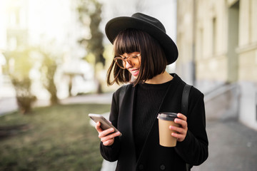 Young pretty woman with coffee cup using smartphone walking in city,