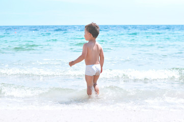 Cute little boy on the beach in white swimming trunks stands in the sea and looks into the distance.